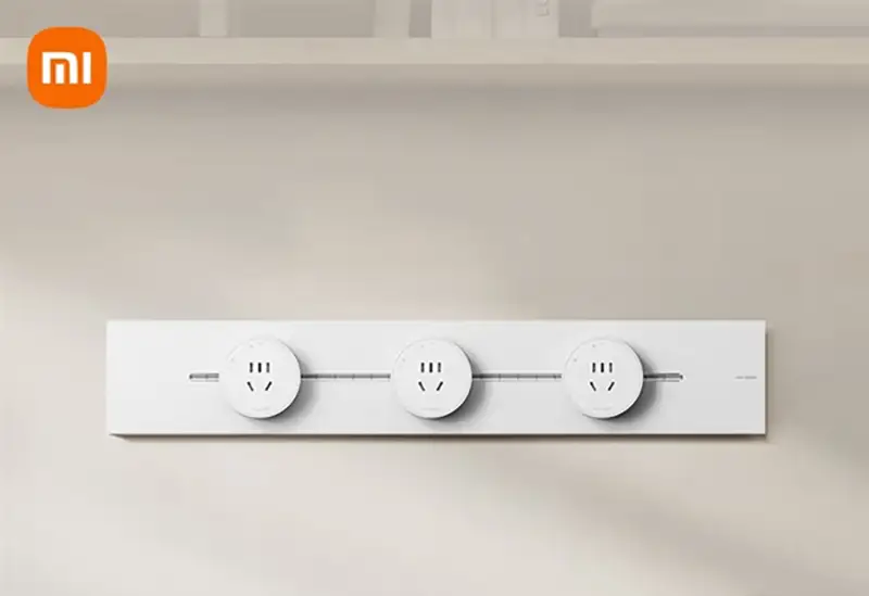 Toma frontal del Xiaomi Track Socket en su versión 'Blanco Nieve', montado horizontalmente en una pared de color crema. El sistema de enchufe modular muestra tres adaptadores redondos distribuidos a lo largo del riel.