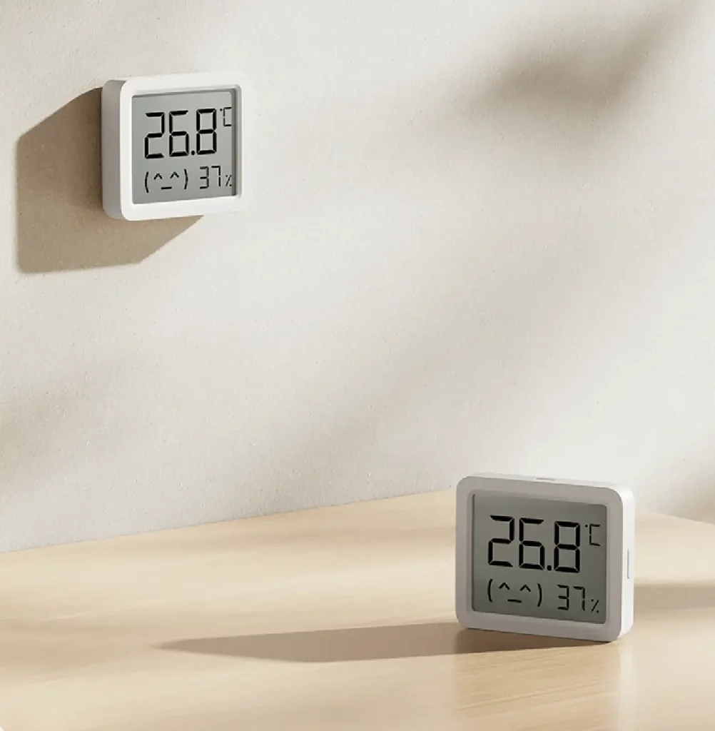 Dos sensores Xiaomi Smart Temperature and Humidity Monitor 3 Mini en un ambiente de hogar. Una unidad está montada en la pared y la otra de pie sobre una mesa de madera, mostrando ambas la temperatura y humedad en su pantalla.