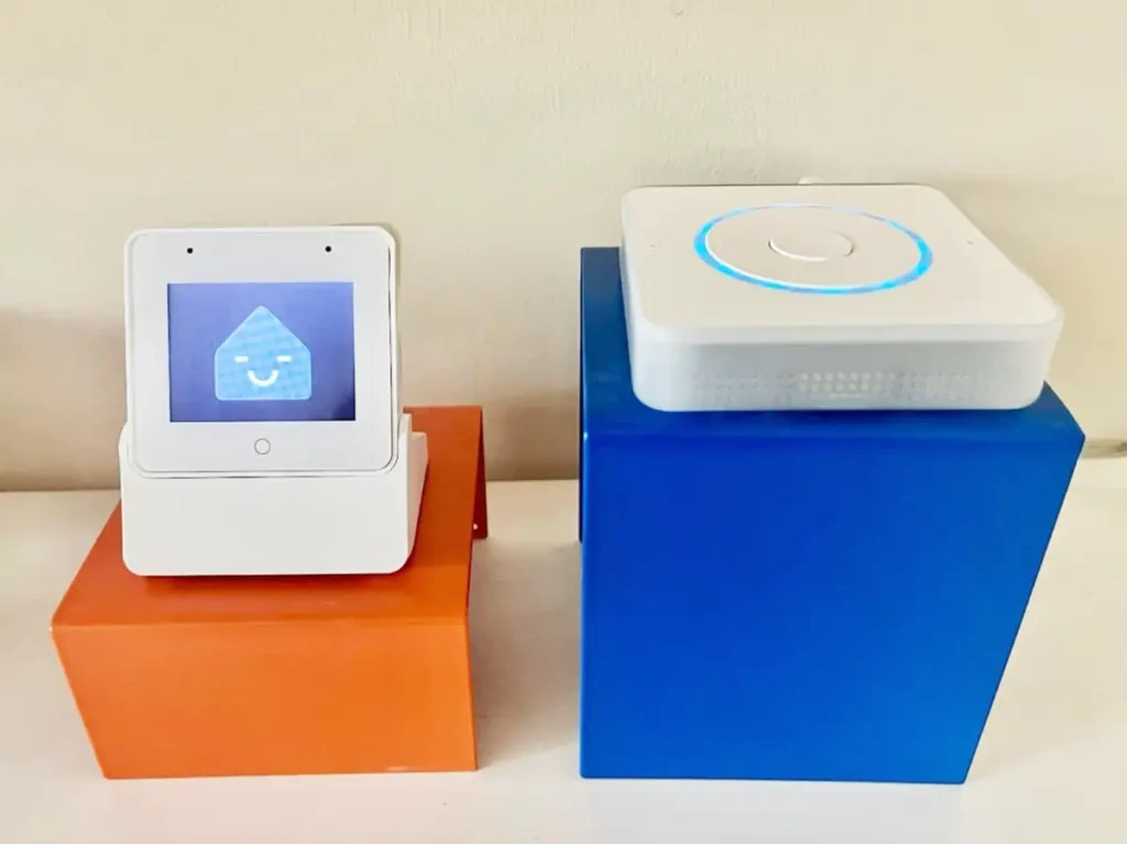 Dos dispositivos de asistente de voz de Home Assistant uno al lado del otro. A la izquierda, el Voice Preview Edition con su pantalla encendida mostrando la mascota Nabu sonriente. A la derecha, el hardware S3 Box 3 con su anillo de luz azul iluminado.