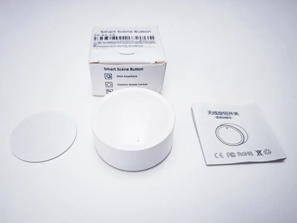 Unboxing de la perilla inteligente Moes/Tuya ZG-101Z/D. La imagen muestra los componentes del producto: el botón-perilla redondo y blanco en el centro, a su izquierda un adhesivo circular, y a su derecha el manual de usuario y la caja de embalaje.