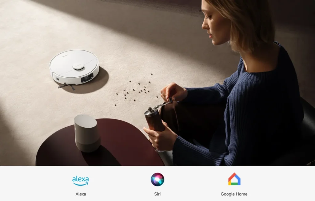 El robot aspirador Dreame D20 Pro Plus en un salón, listo para limpiar unos granos de café derramados. La imagen destaca su compatibilidad con los asistentes de voz Alexa, Siri y Google Home para un control manos libres.