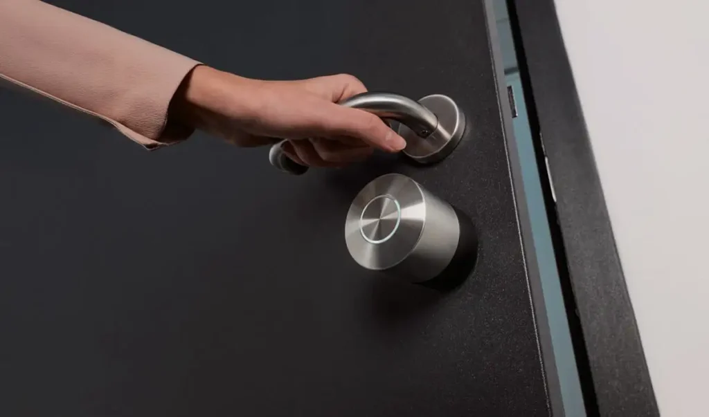 Cerradura inteligente Nuki instalada en una puerta oscura. Una mano está girando la manivela de la puerta, situada justo encima de la cerradura.