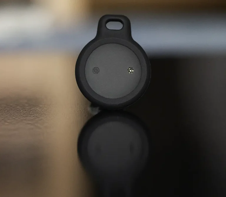 Foto de producto profesional del WAFTING Tracker Tag negro dentro de su funda de silicona, colocado sobre una superficie oscura y brillante que crea un reflejo claro del dispositivo.