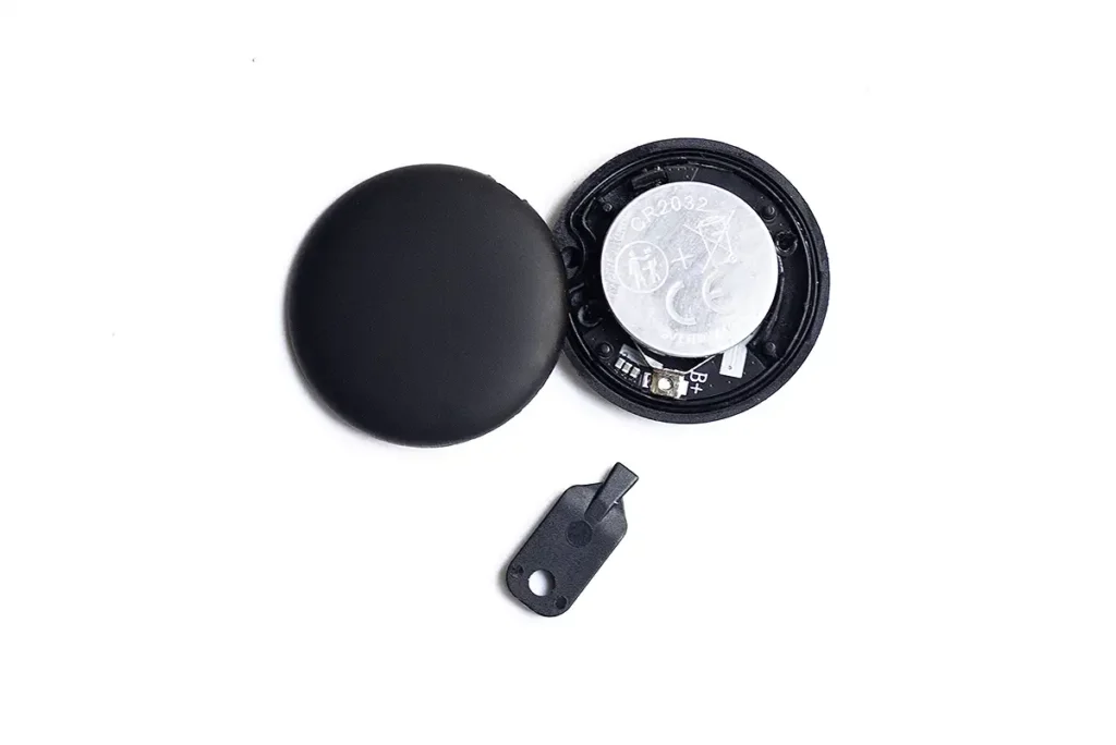 Despiece del localizador Bluetooth WAFTING Tracker Tag sobre un fondo blanco. Se muestra el interior del dispositivo con la pila de botón CR2032 visible, la tapa y la herramienta de apertura, revelando la falta de una junta de estanqueidad para la resistencia al agua