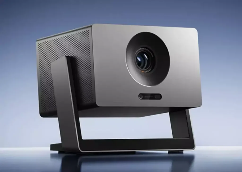 El proyector Xiaomi Laser Projector 3 en una imagen de producto. El dispositivo, de color gris metalizado y diseño cúbico, está inclinado sobre su base-cardán negra integrada, mostrando la lente circular en el frontal y los sensores para el autoenfoque.
