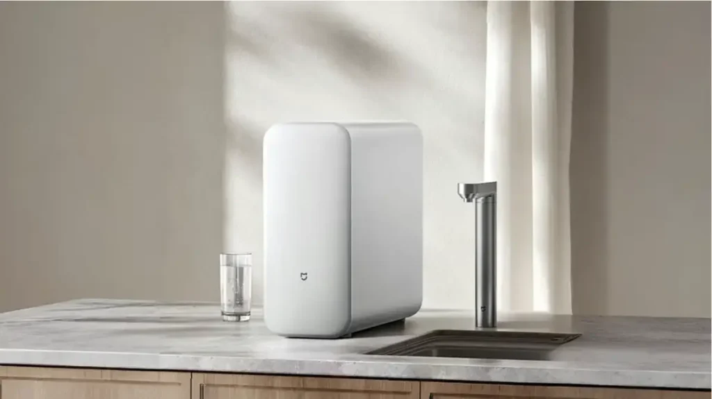 Imagen de ambiente del purificador de agua Xiaomi Mijia S1 1200G instalado en la encimera de una cocina moderna, mostrando su diseño minimalista junto al grifo y un vaso de agua.
