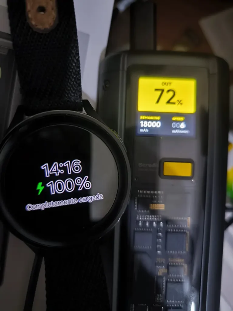 Prueba de carga de un accesorio de bajo consumo: un reloj inteligente en primer plano muestra '100% Completamente cargada', mientras que al lado, la pantalla de la Xiaomi Power Bank indica que tiene un 72% de batería restante (18000 mAh) y está suministrando energía a baja velocidad (006 mAh/min)
