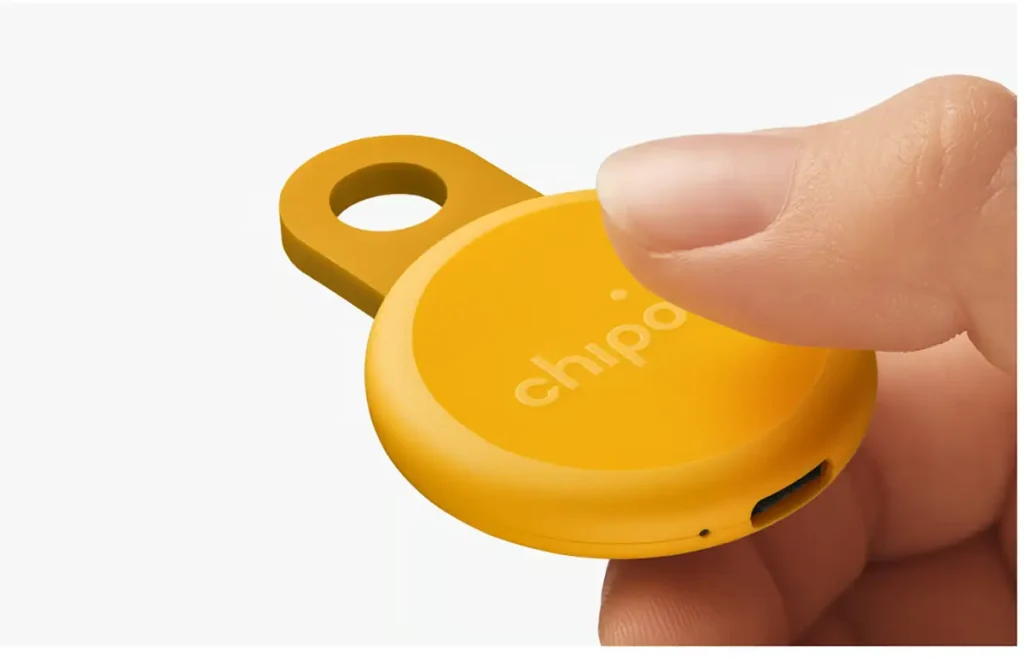 Primer plano del localizador recargable Chipolo Loop de color amarillo, mostrando el puerto de carga USB-C y el aro de silicona para llaves mientras un dedo presiona su botón central.