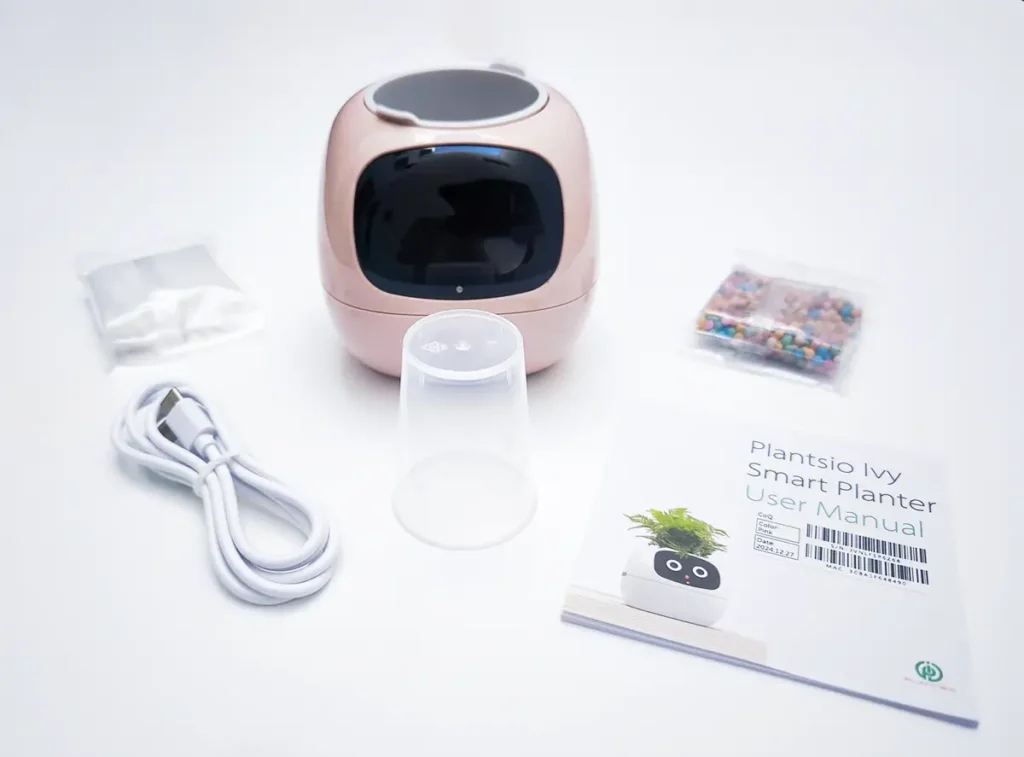 Contenido de la caja del macetero inteligente Ivy Smart Planter. La imagen muestra el dispositivo en color rosa, el cable de carga USB-C, el manual de usuario, el vaso de riego y las bolsas con piedras decorativas blancas y de colores.
