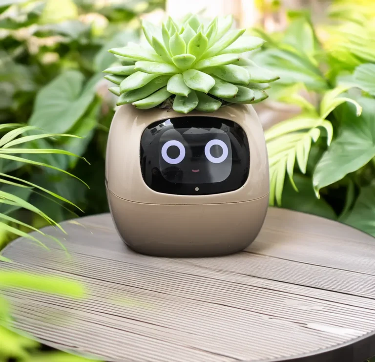 El macetero inteligente Ivy Smart Planter con una planta suculenta, mostrando una cara animada en su pantalla. Está colocado en una mesa de exterior con un fondo de jardín verde y frondoso.