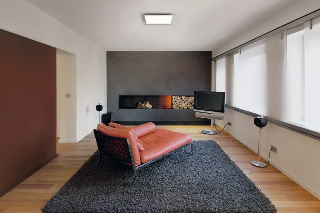Panel LED de techo Osram SMART+ WiFi Planon Plus 450x450 TW, ajustado a 4000K, iluminando un salón de diseño moderno con chimenea, suelo de madera y un sofá de cuero naranja.