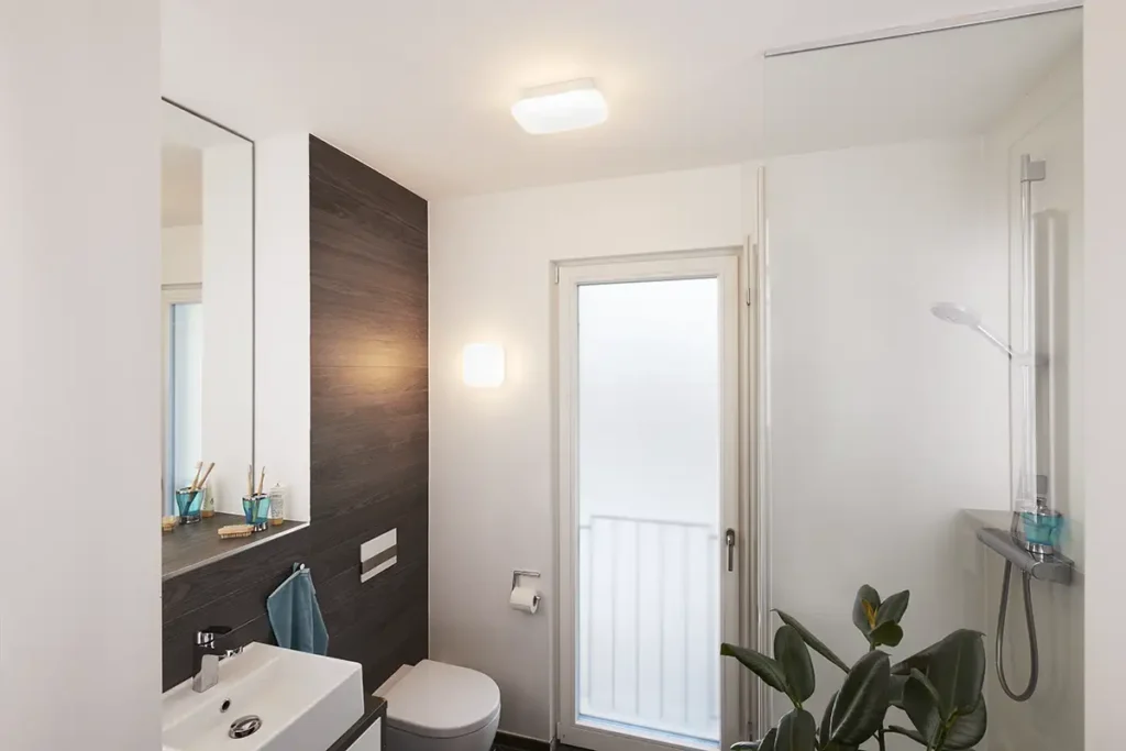 Baño moderno y luminoso con dos luminarias Osram SMART+ Matter Orbis Wall Aqua IP44. Una instalada como plafón de techo y otra como aplique de pared sobre una pared de acento de madera, demostrando su versatilidad y diseño compacto.