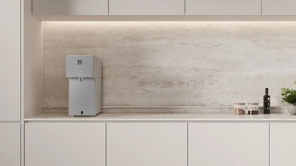 Purificador de agua Samsung Bespoke AI Water Purifier Countertop en color gris/plateado, colocado sobre una encimera de cocina moderna y minimalista con armarios blancos y backsplash de piedra.