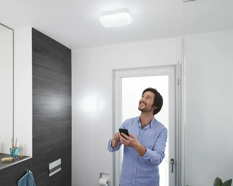 Hombre controlando con su smartphone las luminarias inteligentes Osram SMART+ Matter Orbis Aqua IP44 en un baño moderno. Una lámpara está instalada como plafón de techo y otra como aplique de pared, demostrando el control de la iluminación inteligente.