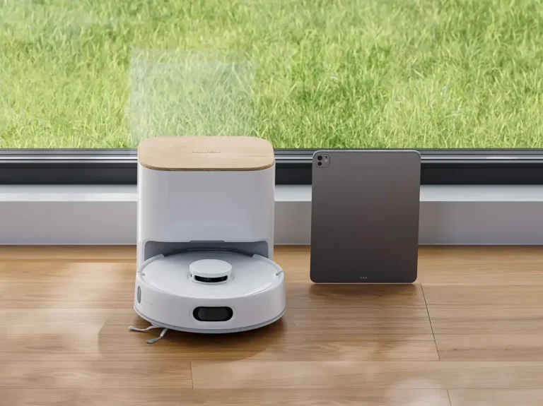 La base de autovaciado del robot SwitchBot K11+ tiene un diseño compacto que ahorra espacio. En la imagen se muestra junto a una tablet para resaltar su reducido tamaño y su estética moderna con tapa de madera.
