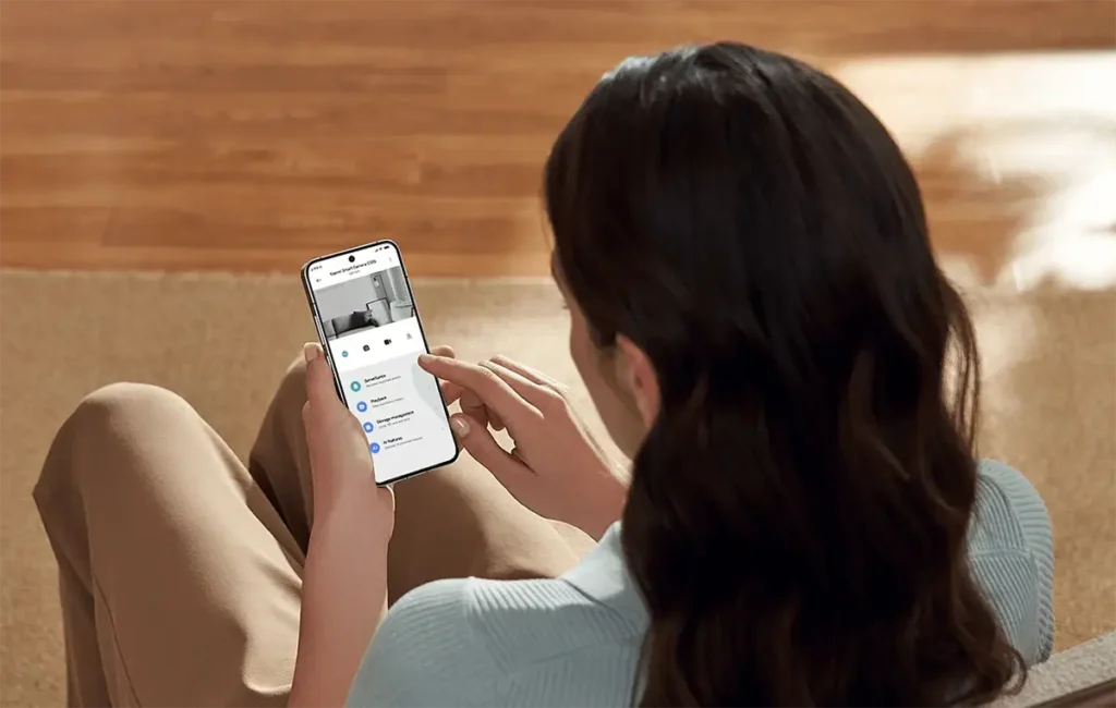 Una persona utilizando la aplicación Xiaomi Home en su smartphone para controlar la Xiaomi Smart Camera C100. En la pantalla se puede ver la transmisión de vídeo en vivo y el menú de ajustes para gestionar las funciones de IA y el almacenamiento.