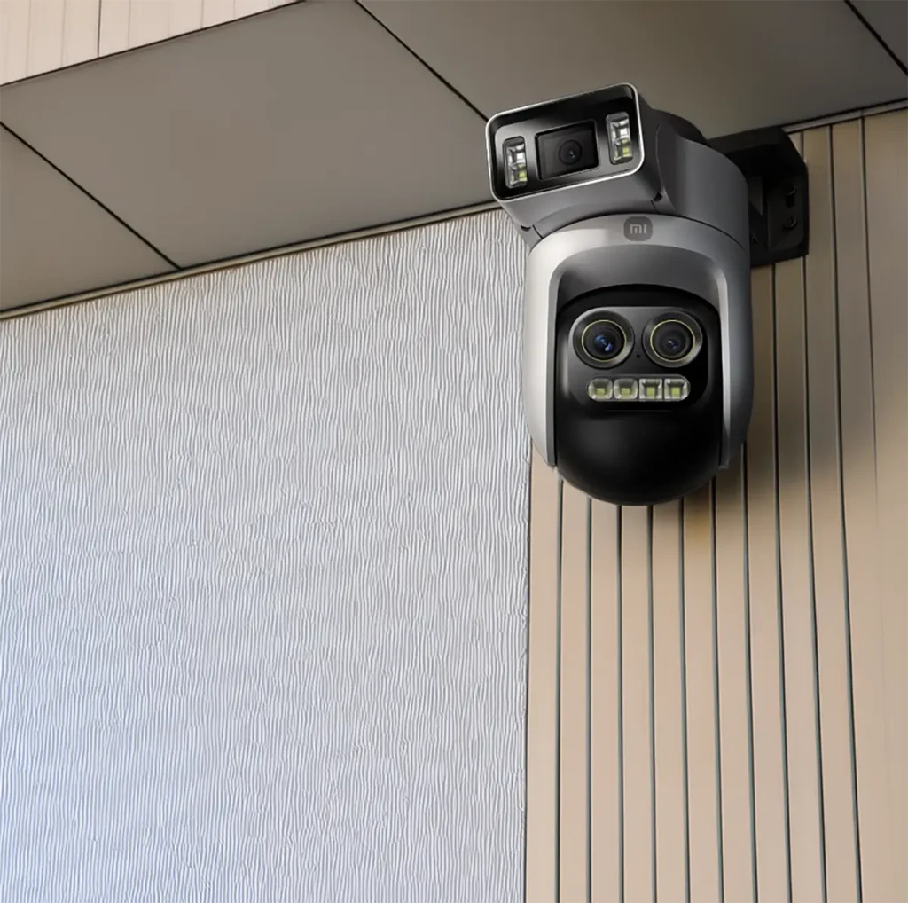 Ilustracion de la cámara Xiaomi Smart Outdoor Camera 4 Pro en un entorno exterior, montada en la esquina de una pared con revestimiento de dos texturas (una blanca ondulada y otra beige con listones verticales). La cámara vigila el exterior del edificio, demostrando su diseño y funcionalidad.