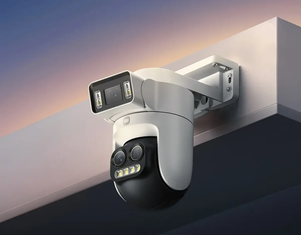 La cámara de seguridad para exteriores Xiaomi Smart Outdoor Camera 4 Pro montada en una pared, mostrando su diseño de triple lente con un cabezal motorizado, y un cielo al atardecer de fondo.