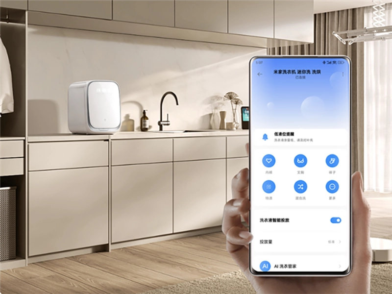 Control remoto de la lavadora-secadora Xiaomi Mijia Mini Wash a través de la aplicación Mi Home en un smartphone, con la máquina instalada al fondo en un lavadero moderno y organizado.