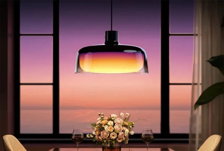 Lámpara colgante inteligente Govee Pendant Light (H60C1) creando una iluminación ambiental sobre una mesa de comedor. La luminaria de vidrio muestra un espectacular degradado de color en tonos cálidos (púrpura, naranja y amarillo), a juego con el atardecer que se ve por la ventana. Escena ideal para mostrar las capacidades de la luz multicolor y el control por zonas.