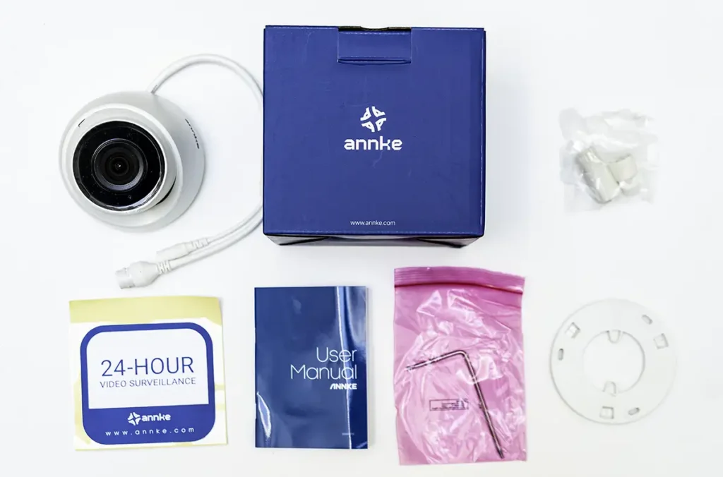 Unboxing completo de la cámara de videovigilancia ANNKE C500 (modelo I51DM01) mostrando todos sus accesorios sobre un fondo blanco: la cámara tipo torreta, la caja del producto, el manual, la llave Allen, los tornillos y tacos de montaje, y la pegatina de zona videovigilada.