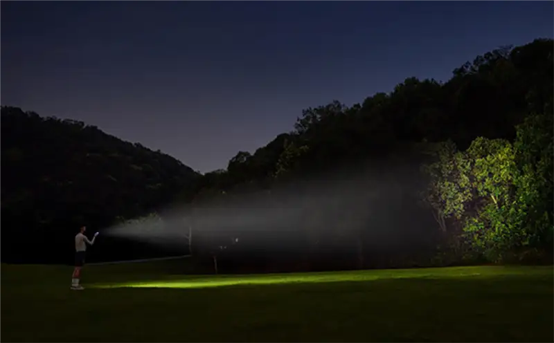Demostración de la potencia de la linterna Xiaomi Mijia Outdoor Multifunctional Light de 600 lúmenes por la noche. Una persona en un campo ilumina un bosque lejano, mostrando el impresionante alcance de 110 metros del haz de luz, ideal para camping y senderismo.
