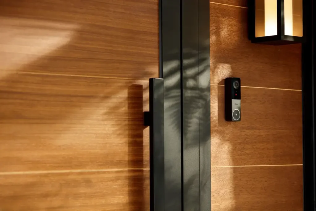 El nuevo videotimbre Philips Hue Secure instalado en la pared de madera de una entrada moderna. El timbre inteligente, parte del ecosistema de seguridad para el hogar de Hue, se integra perfectamente con el diseño gracias a su acabado en negro y plata. La imagen muestra el dispositivo junto a una puerta y una lámpara de exterior, destacando su estética elegante para un hogar conectado.
