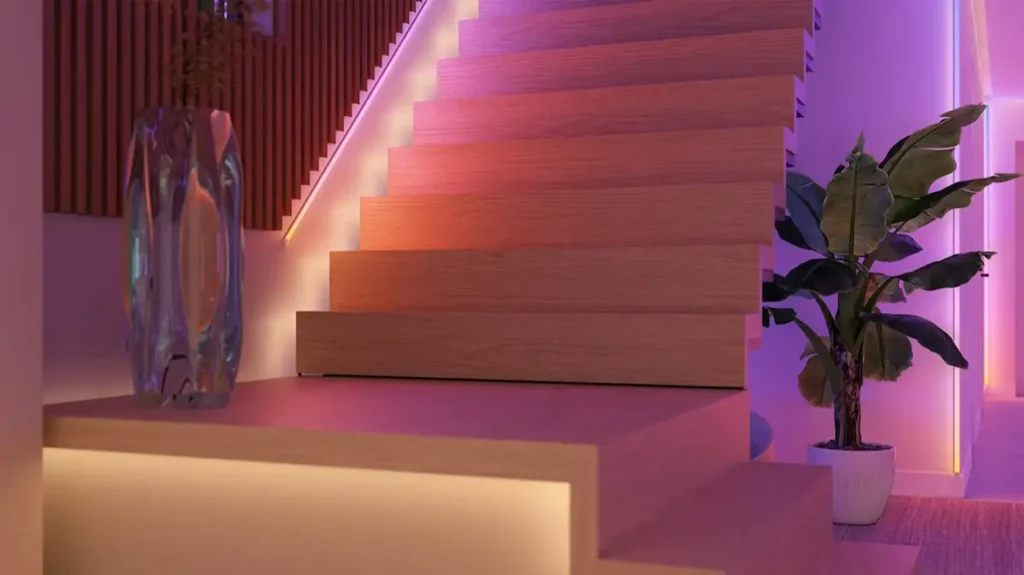 Las nuevas tiras de luces LED Philips Hue iluminan una moderna escalera de madera, creando un degradado de color ambiental perfecto. La escena muestra la transición suave de tonos naranja cálido a rosa y púrpura, destacando la versatilidad de los nuevos lightstrips de Hue para el diseño de interiores y la decoración de un hogar inteligente.