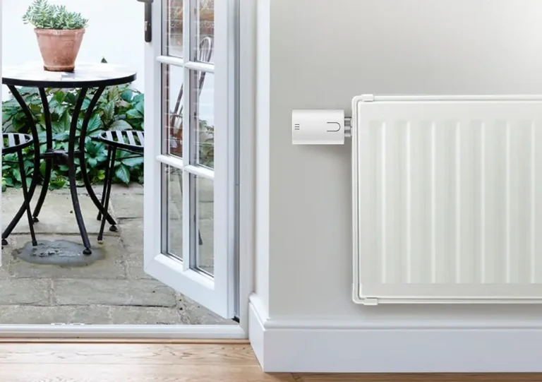 Cabezal termostático inteligente Meross MTS150 con Matter instalado en un radiador blanco. La imagen ilustra la función de detección de ventana abierta, mostrando una puerta de patio abierta al fondo para un control eficiente de la calefacción.