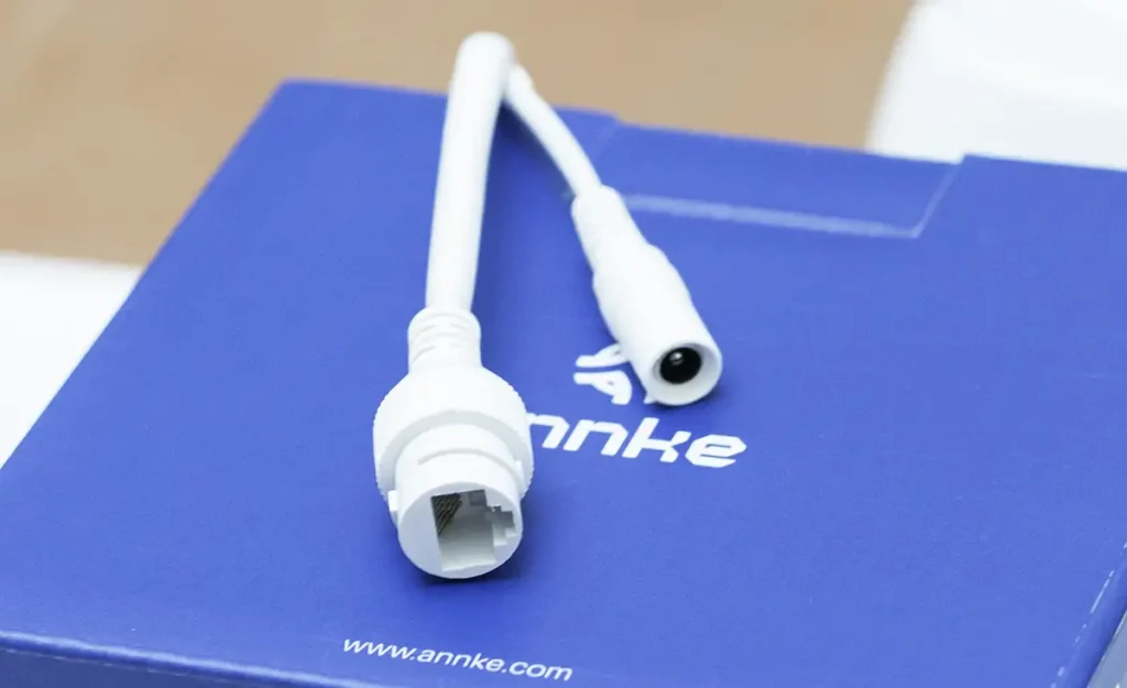 Primer plano de los conectores de la cámara de seguridad ANNKE. Se muestra el puerto de red Ethernet RJ45 para alimentación PoE y datos, y la entrada de corriente alternativa de 12V DC, ambos de color blanco sobre la caja azul de la marca