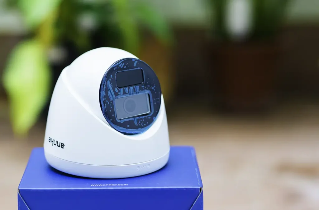 Cámara de seguridad PoE ANNKE C300 tipo domo, de color blanco, se muestra en un primer plano sobre su caja azul con un fondo desenfocado de plantas