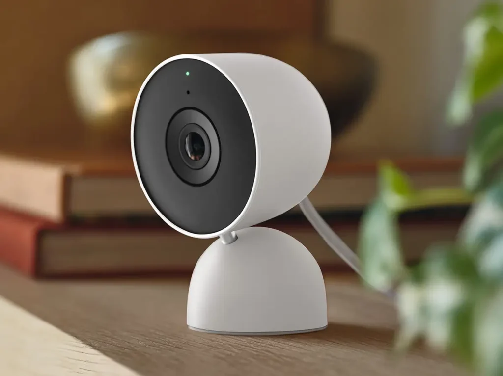 La nueva cámara de seguridad Google Nest Cam Interior 2K (3ª generación) en color blanco, colocada sobre una estantería de madera en el interior de una casa, mostrando su base y el objetivo de la cámara.