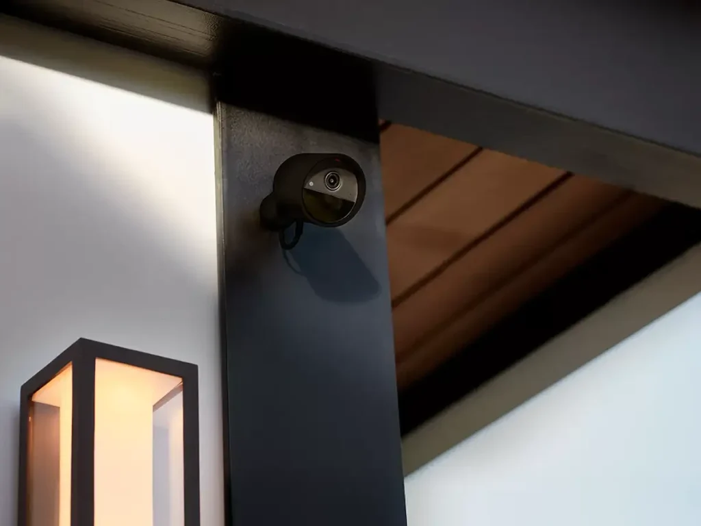 Cámara de seguridad Philips Hue Secure 2K con cable en color negro, instalada en el exterior de una casa para la vigilancia del hogar. Al lado, un aplique de luz moderno muestra la integración con el ecosistema de iluminación inteligente