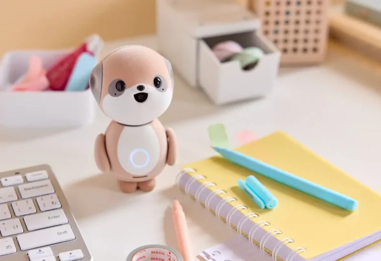 El robot Sharp Poketomo, con un adorable diseño de perro en tonos marrones, está sentado sobre un escritorio blanco junto a un teclado, un cuaderno amarillo y varios útiles de oficina.
