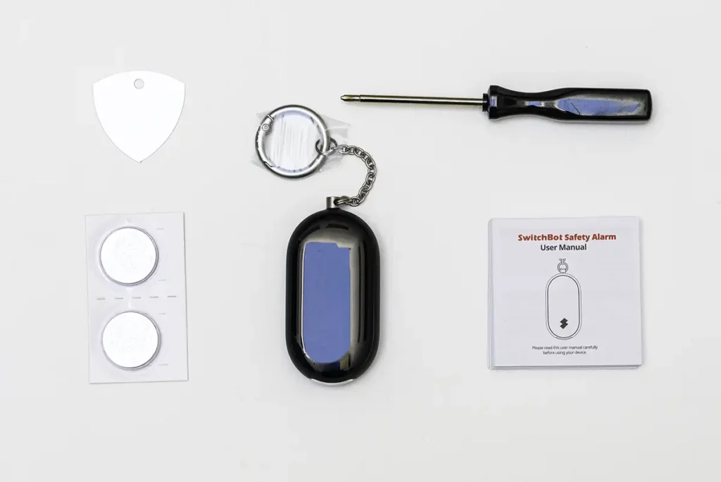 Unboxing del SwitchBot Safety Alarm con todos sus componentes dispuestos sobre una superficie blanca. Se ve el dispositivo de alarma personal de color negro con su llavero, dos pilas de botón CR2032, un destornillador de precisión, el manual de usuario y una púa blanca para abrir el compartimento.