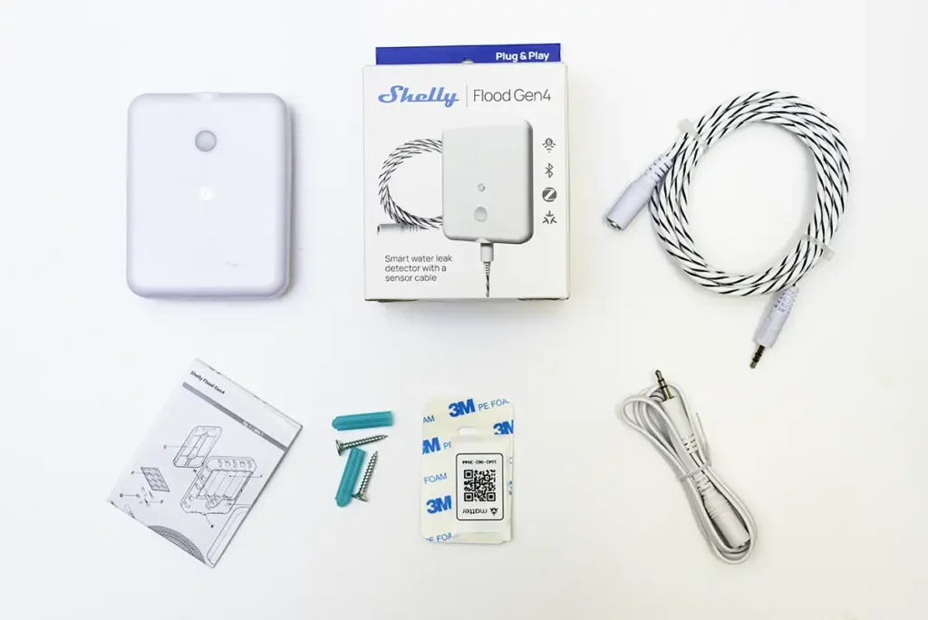 Unboxing completo del sensor de fugas de agua Shelly Flood Gen4. Se muestra todo el contenido de la caja: el dispositivo principal, el cable detector trenzado de 2 metros, un extensor, el manual de usuario y el kit de montaje con tornillos, tacos y una pegatina adhesiva 3M con el código QR para la configuración de Matter.