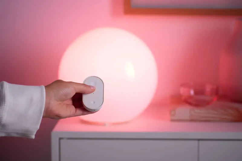 Mando a distancia BILRESA de IKEA controlando una lámpara inteligente compatible con Matter, cambiando la luz a un tono rosado para el nuevo ecosistema de hogar inteligente.