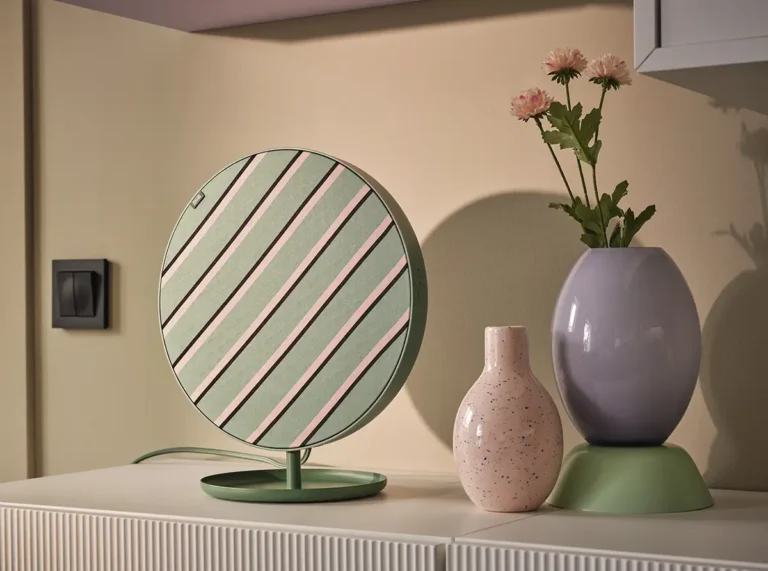 Altavoz circular IKEA Solskydd de diseño en verde y rosa sobre mueble de salón, acompañado de jarrones y flores como parte de la decoración.