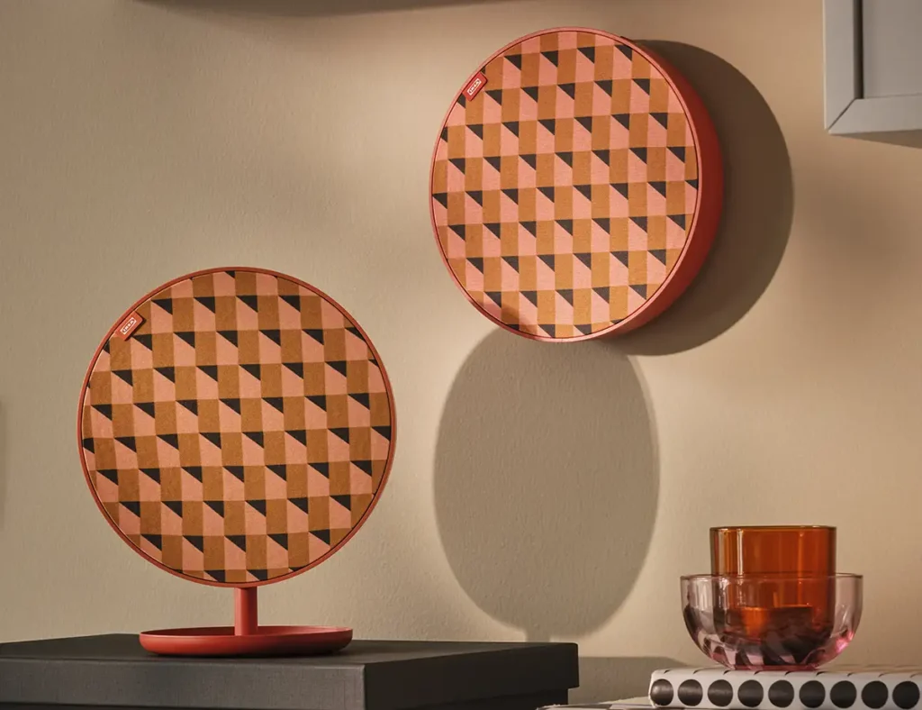 Dos altavoces Bluetooth IKEA Solskydd en color naranja con patrón geométrico, uno sobre peana y otro colgado en la pared junto a vasos de cristal.