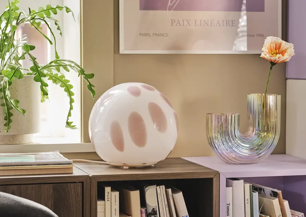 Detalle de la lámpara de mesa redonda IKEA FADO con patrón rosa, situada sobre mueble con libros y accesorios decorativos.