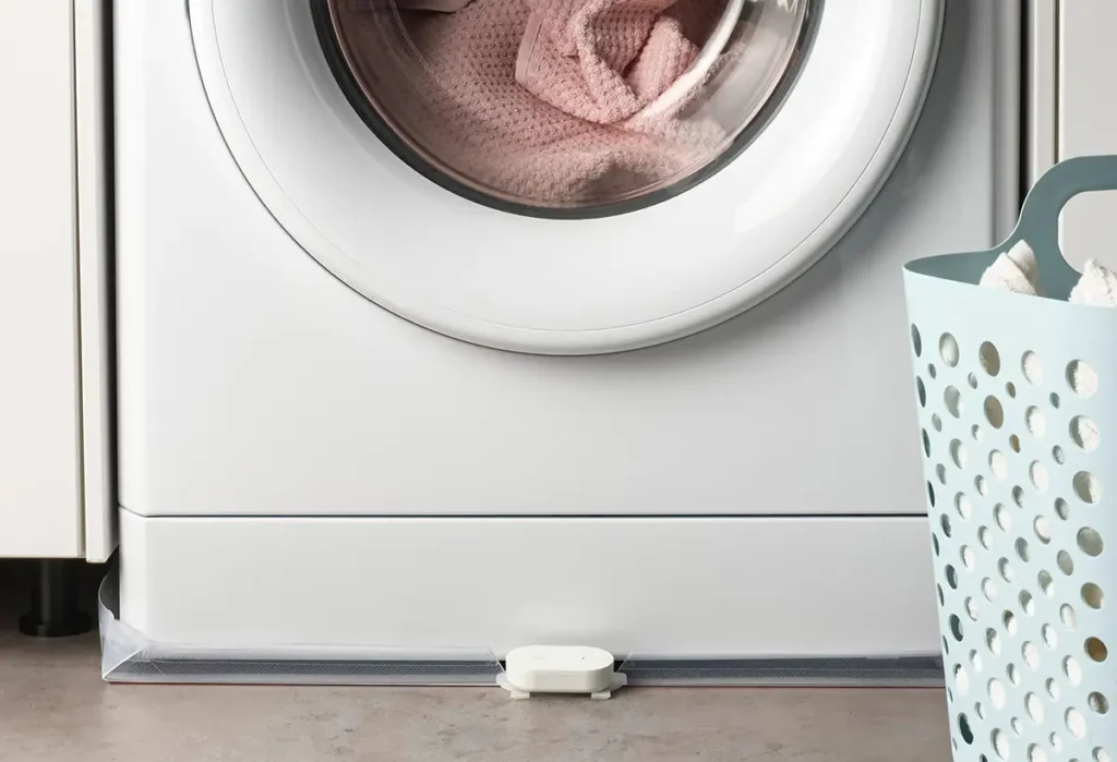 El nuevo sensor de fugas de agua IKEA KLIPPBOK colocado en el suelo frente a una lavadora para detectar escapes y prevenir daños. Este dispositivo inteligente compatible con Matter envía notificaciones al móvil.