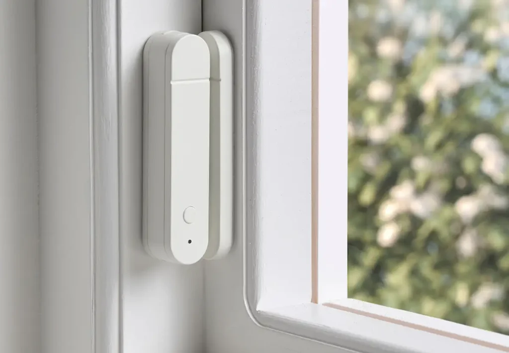 El nuevo sensor de puerta y ventana IKEA MYGGBETT instalado en el marco de una ventana blanca, compatible con Matter para enviar notificaciones y activar automatizaciones en el hogar inteligente.