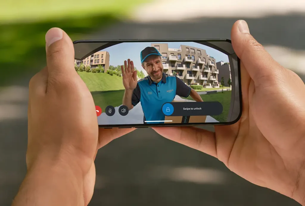 Una persona usando la app de Ring en su móvil para ver a un repartidor de Amazon que ha llamado al portal. La pantalla muestra la opción de deslizar para desbloquear ("Swipe to unlock") y abrir la puerta a distancia.