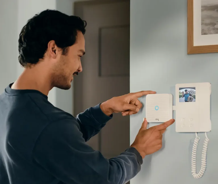 Un hombre instalando el nuevo Ring Intercom Video junto a un videoportero tradicional. La pantalla del interfono muestra a un repartidor con un paquete, ilustrando cómo el dispositivo permite ver a las visitas de forma remota.