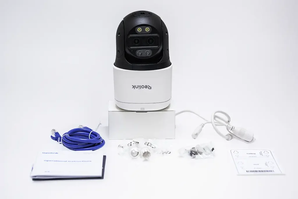 Unboxing de la cámara de seguridad Reolink TrackMix PoE: cuerpo de la cámara PTZ 4K de doble lente sobre su base, acompañada por cable Ethernet azul, manuales, cable de conexión PoE, bolsa con tornillos y tacos de montaje y plantilla para fijarla en techo o pared, sobre fondo blanco.