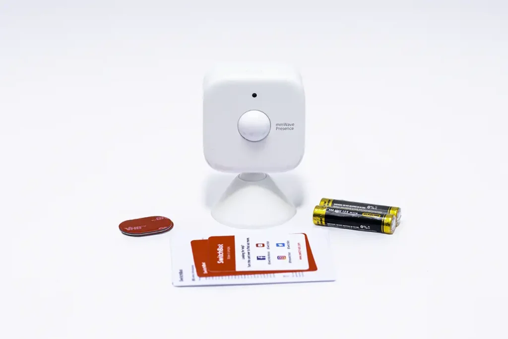 Contenido de la caja del SwitchBot Presence Sensor: sensor mmWave con base magnética, dos pilas AAA incluidas, adhesivos 3M y documentación sobre fondo blanco