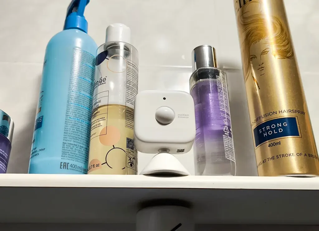 Sensor de presencia SwitchBot mmWave colocado en una balda del baño entre botes de cosmética y laca, aprovechando su protección IP55 para automatizar la luz en zonas húmedas