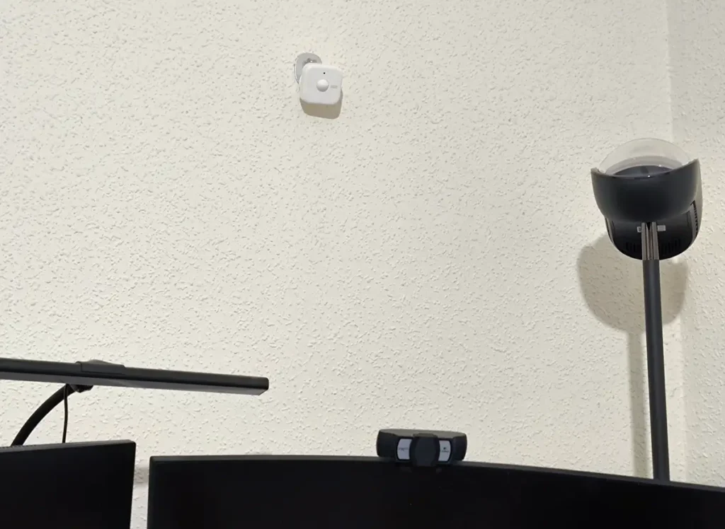 Sensor de presencia SwitchBot montado en la pared sobre el escritorio, junto a una lámpara de pie y el monitor del ordenador, para detectar ocupación en la habitación de trabajo