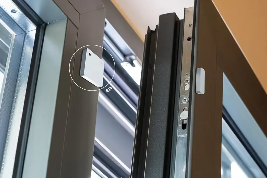 Sensor Shelly Wave Door/Window LR instalado en una puerta corredera de aluminio, mostrando el contacto magnético para detectar la apertura y cierre.