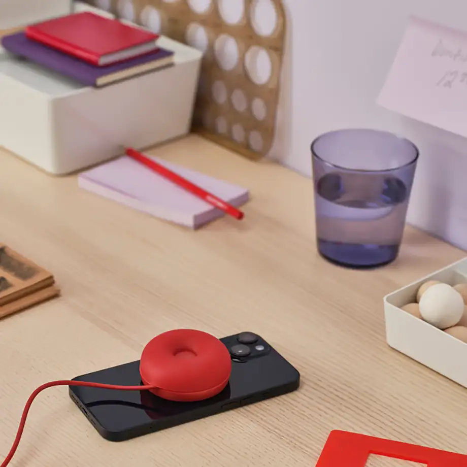 Base de carga IKEA VÄSTMÄRKE en rojo con forma de donut, conectada por cable y cargando un móvil sobre una mesa de escritorio.